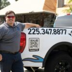 Smiling businessman in gray polo shirt standing next to white work truck with phone number